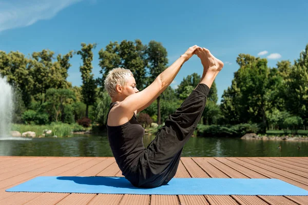 Vista lateral de la mujer practicando yoga y estirando las piernas en la esterilla de yoga cerca del río en el parque - foto de stock