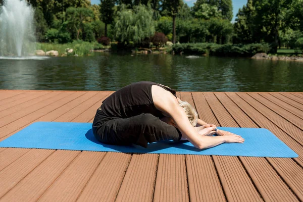 Vista lateral de la mujer practicando yoga en pose Wide Child (Balasana) sobre colchoneta de yoga cerca del río en parque - foto de stock