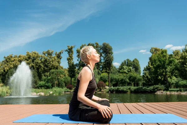 Vista lateral de la mujer feliz practicando yoga en pose de loto en esterilla de yoga cerca del río en el parque - foto de stock