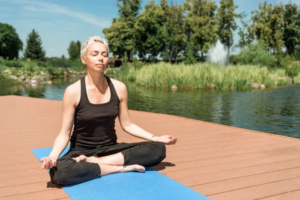 Woman practicing yoga in lotus pose and meditating on yoga mat near river in park — Stock Photo