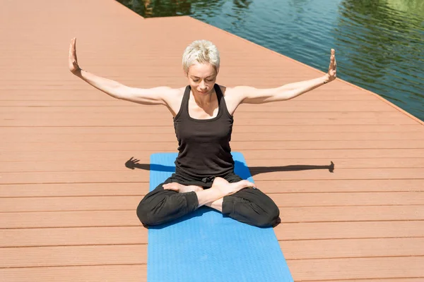 Woman practicing yoga in lotus pose with open arms near river in park — Stock Photo