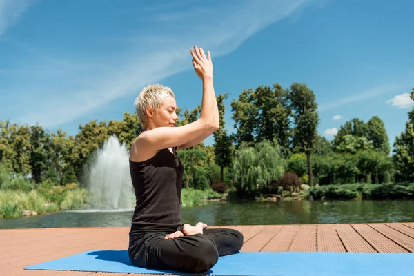 Vista lateral de la mujer practicando yoga en pose de loto y manos en gesto namaste en esterilla de yoga cerca del río en parque - foto de stock