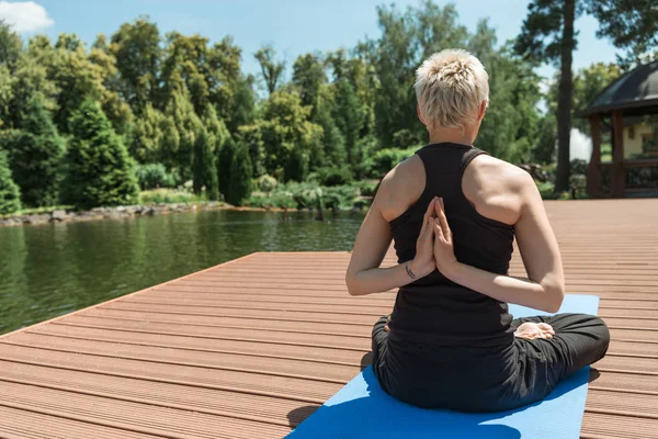 Vue arrière de la femme pratiquant le yoga en pose de lotus et les mains en geste namaste sur tapis de yoga près de la rivière dans le parc — Photo de stock