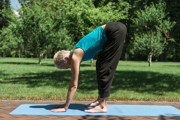 Mujer practicando yoga y haciendo pose de elevación a medio camino (Ardha Uttanasana) sobre estera de yoga en parque - foto de stock