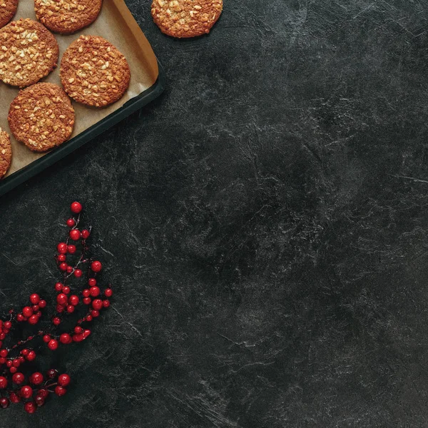 Vista superior de biscoitos recém-assados na bandeja e bagas vermelhas na superfície de concreto preto — Fotografia de Stock