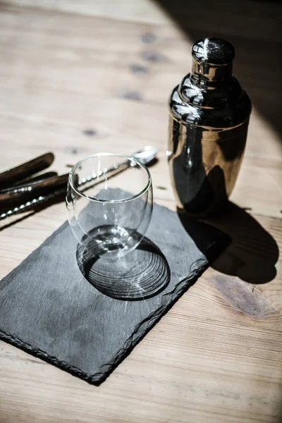 Крупным планом глядя на блестящий пустой стакан на доске, шейкер и щипцы на деревянном столе — стоковое фото