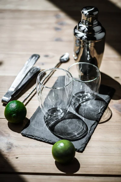 Крупный план свежих лаймов, пустых стаканов, щипцов и шейкера на деревянном столе — стоковое фото