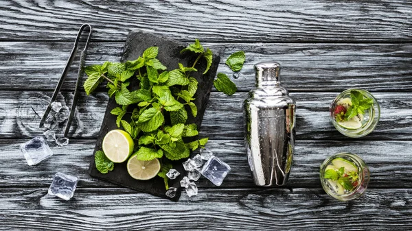 Vue de dessus de menthe fraîche, pinces, shaker et fraise kiwi mojito dans des verres sur table en bois — Photo de stock