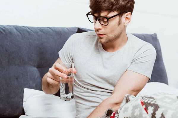 Selektiver Fokus eines jungen Mannes in Brille, der zu Hause im Bett Wasser trinkt — Stockfoto