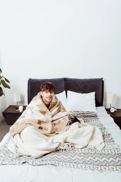 Blick aus der Vogelperspektive auf einen kranken jungen Mann, der in eine Decke gehüllt zu Hause auf dem Bett sitzt — Stockfoto