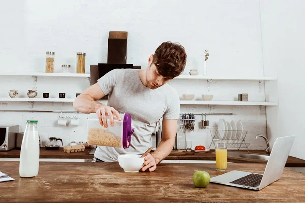 Freelancer masculino fazendo café da manhã com flocos de milho à mesa com laptop na cozinha em casa — Fotografia de Stock