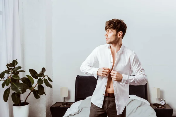 Side view of handsome businessman buttoning up white shirt in bedroom at home — Stock Photo