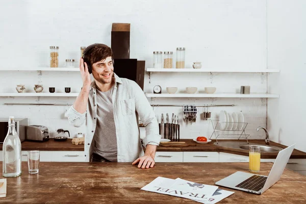 Feliz hombre freelancer en los auriculares de pie cerca de la mesa con el ordenador portátil en la cocina en casa - foto de stock