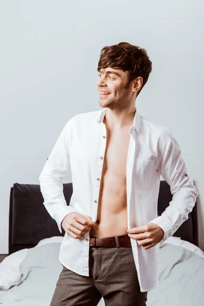 Sonriente joven hombre de negocios abotonando camisa blanca en el dormitorio en casa - foto de stock