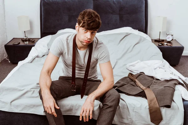 Уставший молодой бизнесмен с поясом на шее смотрит в камеру и сидит на кровати дома — стоковое фото