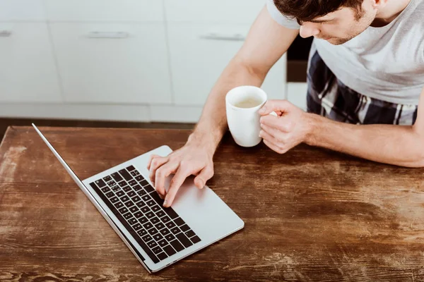 Vista ad alto angolo di freelance maschio con tazza di caffè che lavora su computer portatile a tavola in cucina — Foto stock