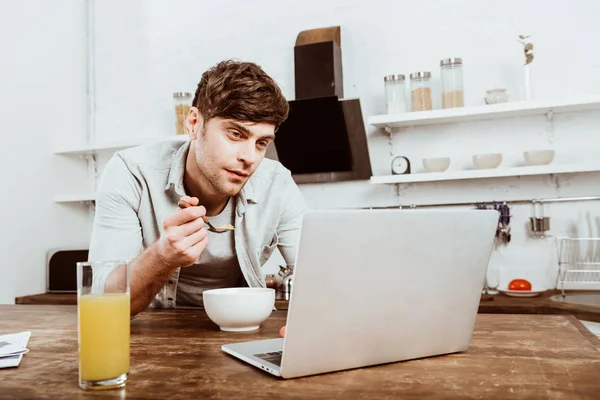 Freelance mâle manger des flocons de maïs et de travailler sur ordinateur portable à la table dans la cuisine — Photo de stock