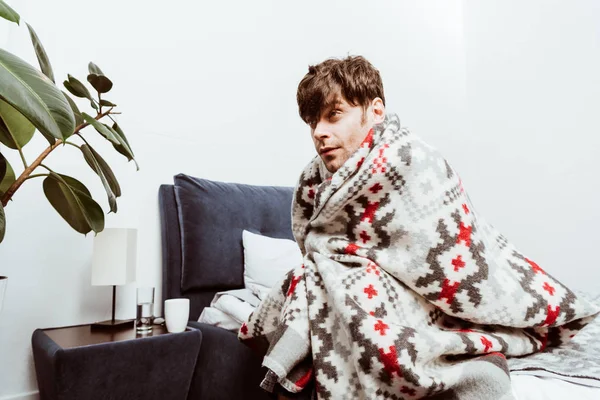 Вибірковий фокус хворого молодого чоловіка, загорнутого в ковдру, сидить на ліжку вдома — стокове фото