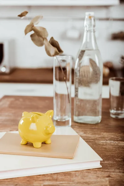 Vue rapprochée de tirelire jaune sur le manuel à table en bois avec bouteille d'eau et vase avec branche — Photo de stock