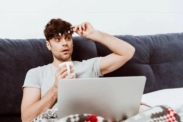 Jovem freelancer masculino decolando óculos e segurando xícara de café na cama com laptop em casa — Fotografia de Stock