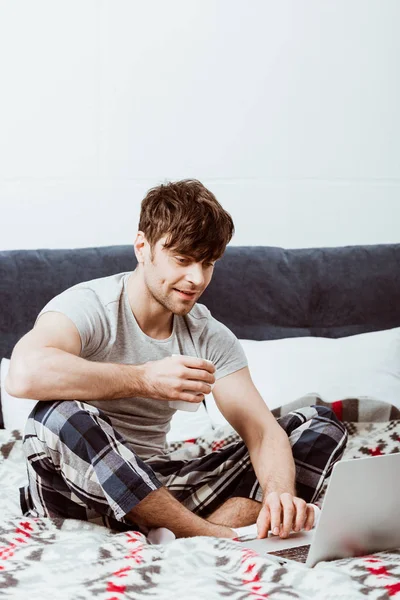Freelancer masculino sorridente com xícara de café sentado na cama com laptop em casa — Fotografia de Stock