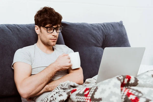 Focused male freelancer in eyeglasses holding coffee cup sitting on bed with laptop at home — Stock Photo