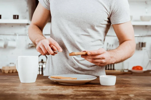 Imagen recortada de hombre extendiendo tostadas por mermelada en la cocina en casa - foto de stock