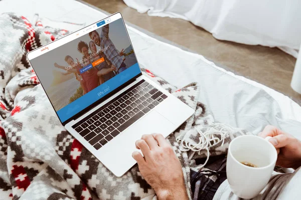 Cropped image of man holding coffee cup and using laptop with couchsurfing on screen in bed at home — Stock Photo
