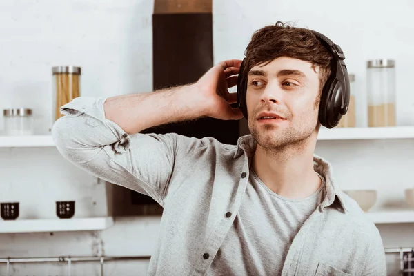 Hombre guapo en auriculares escuchando música y mirando hacia otro lado en la cocina en casa - foto de stock