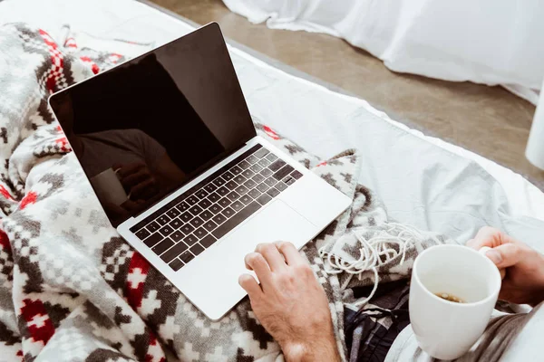 Cropped image of male freelancer with cup of coffee working on laptop in bed at home — Stock Photo