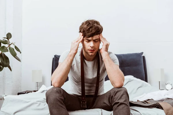 Young businessman with belt over neck sitting on bed and having headache at home — Stock Photo