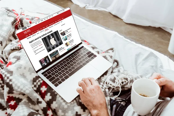 Immagine ritagliata di uomo in possesso di tazza di caffè e utilizzando il computer portatile con bbc notizie sullo schermo a letto a casa — Foto stock
