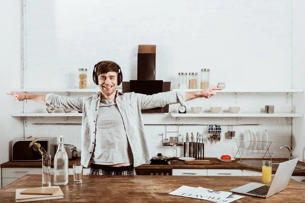 Joyeux freelance masculin dans les écouteurs debout avec de larges bras près de la table avec ordinateur portable dans la cuisine à la maison — Photo de stock