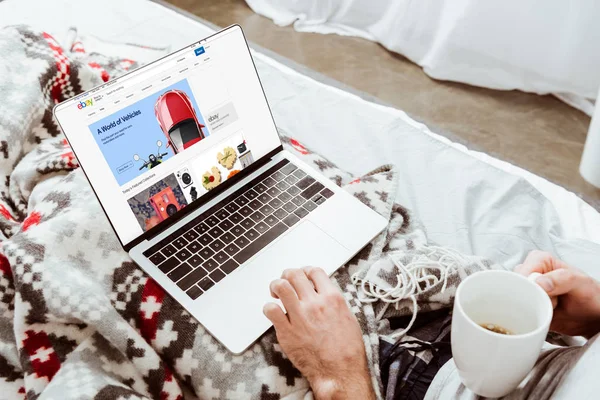 Cropped image of man holding coffee cup and using laptop with ebay on screen in bed at home — Stock Photo