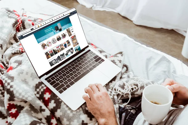 Imagen recortada del hombre sosteniendo la taza de café y el uso de ordenador portátil con Amazon en la pantalla en la cama en casa - foto de stock