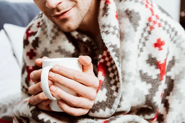Visão parcial do homem doente envolto em cobertor sentado com xícara de chá na cama em casa — Fotografia de Stock