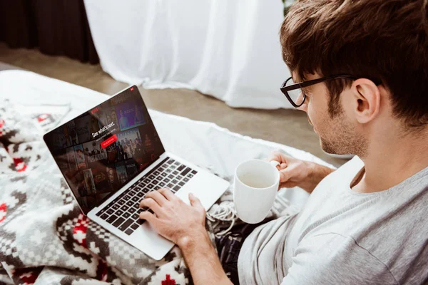 Вид сбоку человека, держащего чашку кофе и использующего ноутбук с Netflix на экране в постели дома — стоковое фото