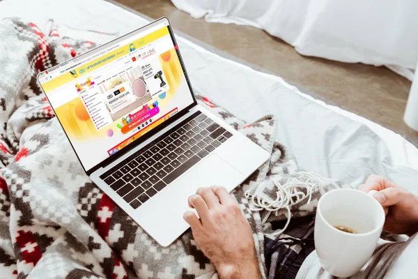 Abgeschnittenes Bild eines Mannes mit Kaffeetasse und Laptop mit aliexpress auf dem Bildschirm im heimischen Bett — Stockfoto