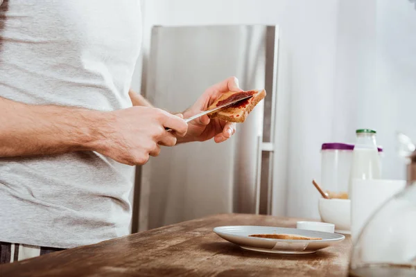 Vista parcial del hombre extendiendo tostadas por mermelada en la cocina en casa - foto de stock