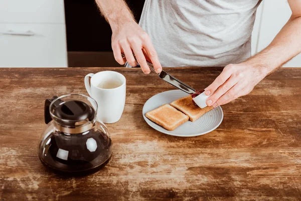 Обрезанный образ человека, распространяющего тост за вареньем за столом с кофейником и чашкой на кухне — стоковое фото