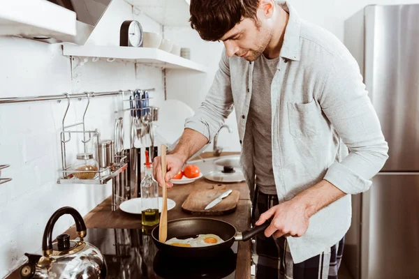 Beau jeune homme faisant des œufs sur le petit déjeuner dans la cuisine à la maison — Photo de stock