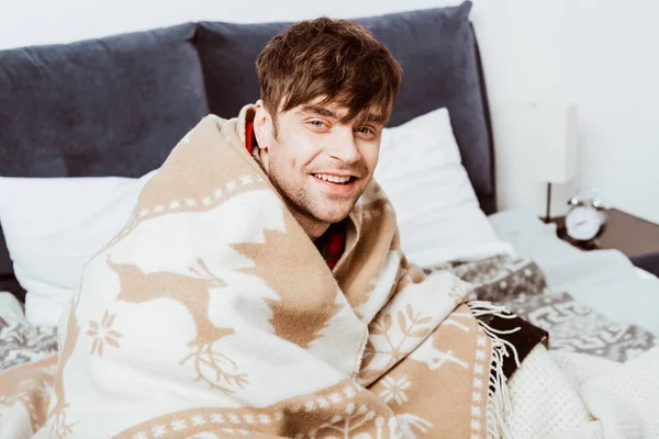 Riéndose joven envuelto en manta sentado en la cama en casa — Stock Photo