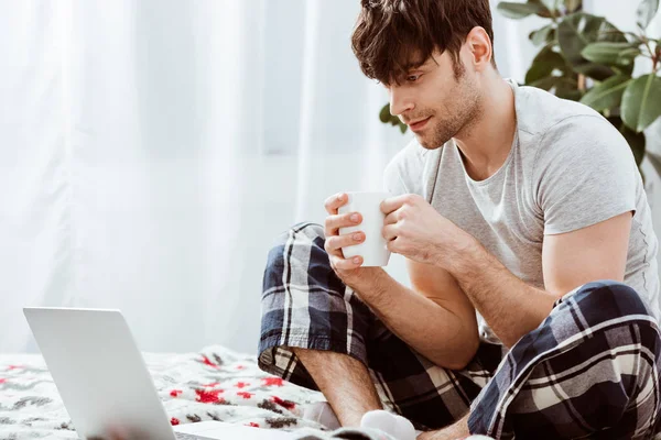 Красивый мужчина фрилансер с чашкой кофе сидит на кровати с ноутбуком дома — стоковое фото