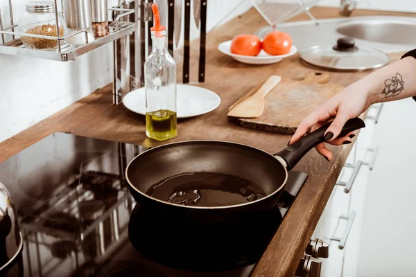 Cropped image of woman with tattooed hand putting frying pan with oil on stove in kitchen at home — Stock Photo
