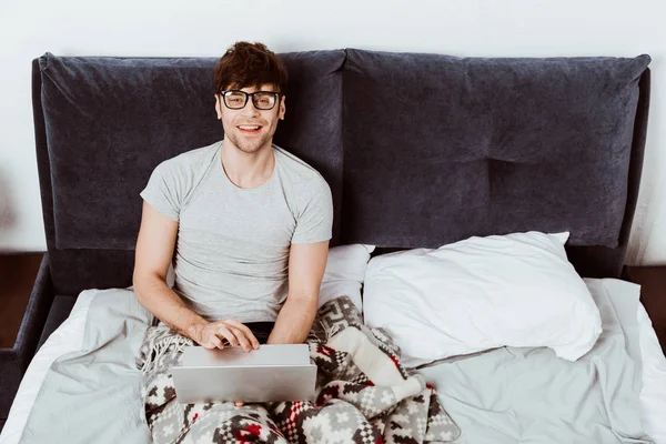 Joyeux mâle pigiste travaillant sur ordinateur portable et regardant la caméra dans le lit à la maison — Photo de stock