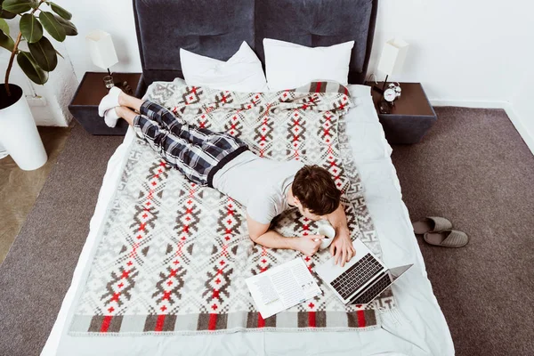 High angle view of male freelancer with coffee cup laying on bed with laptop at home — Stock Photo