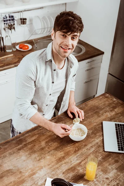 High angle view of male freelancer eating corn flakes on breakfast at table with laptop in kitchen at home — Stock Photo