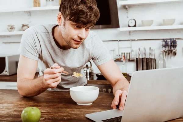 Enfoque selectivo de joven freelancer masculino comiendo hojuelas de maíz en el desayuno en la mesa con el ordenador portátil en la cocina en casa - foto de stock