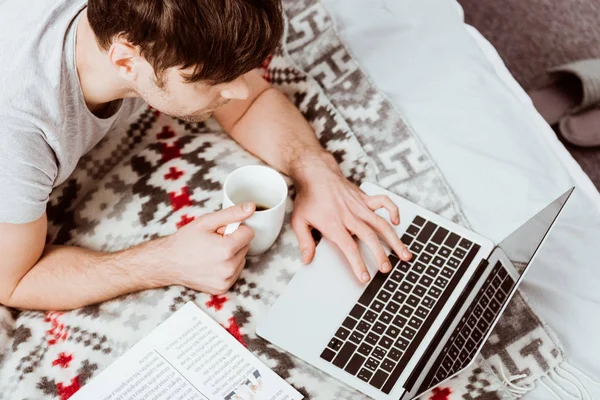 Visão de alto ângulo do freelancer masculino com xícara de café trabalhando no laptop com tela em branco na cama em casa — Fotografia de Stock