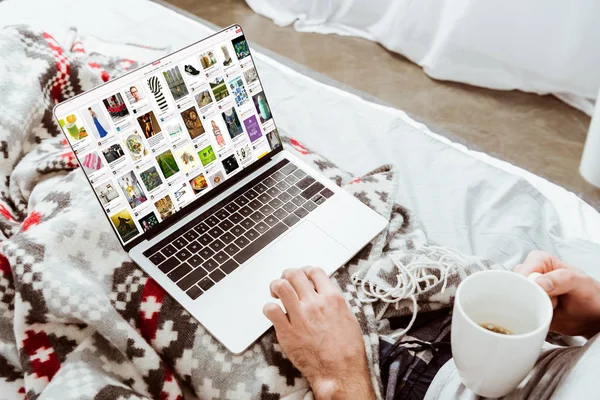 Обітнутого зображення людина тримає чашку кави і використання ноутбука з pinterest на екрані в ліжку у себе вдома — стокове фото